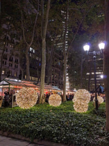 Kerstversiering in Bryant park: grote bollen met lichtjes tussen het groen en kale bomen, op de achtergrond kerststalletjes die er uit zien als serres met warme lichten