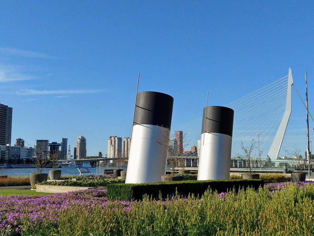 Kunstwerk schoorstenen van grote schepen verwerkt in de tuin op Wilheminapier met zicht op de Nieuwe Maas en de Erasmusbrug in Rotterdam